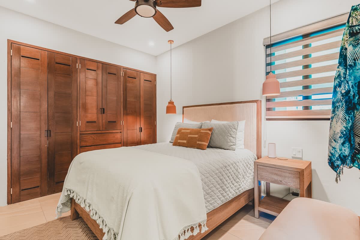 This bedroom features a queen-sized bed with a light beige upholstered headboard, flanked by wooden nightstands with modern orange pendant lights. A wall of dark wooden built-in wardrobes provides ample storage, enhancing the room's clean aesthetic.