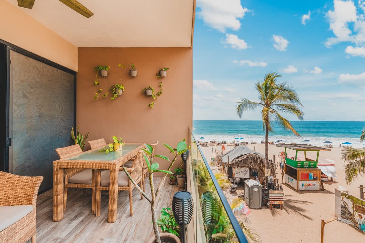 This balcony offers a panoramic view of a sandy beach with a rustic beach bar, the ocean, and palm trees, seen from a glass-railed outdoor dining area with a rattan table and chairs. Hanging and potted plants decorate the warm-toned wall.