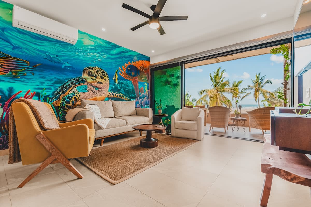 Bright living room with a beige sofa, a yellow armchair, and a white armchair, all facing a vibrant underwater mural, with sliding glass doors opening to a balcony with ocean views and palm trees.