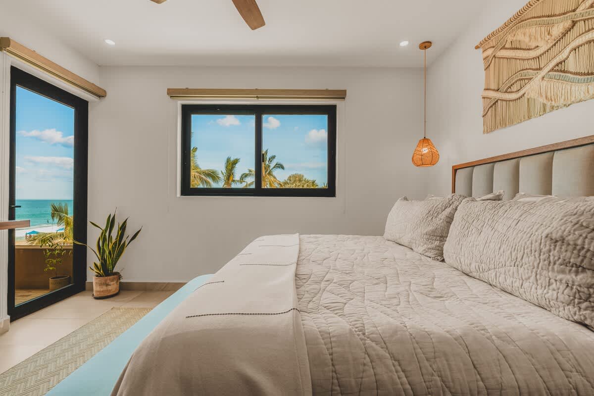 This bedroom offers stunning ocean views through a large window and a glass door leading to a balcony. The room features a large bed with a light duvet and a light blue bench, along with a ceiling fan.