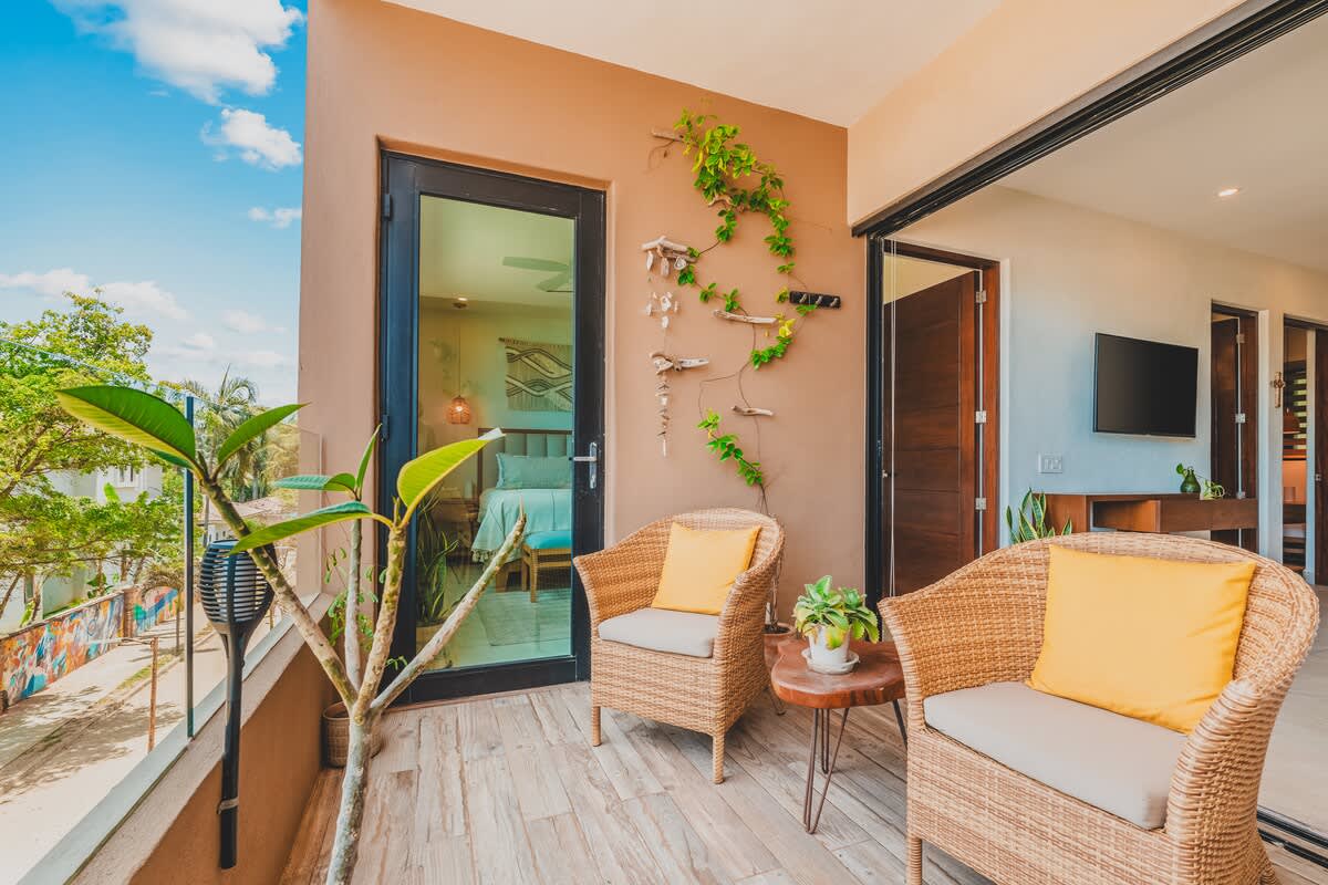 A cozy balcony features two wicker armchairs with yellow cushions and a small wooden side table, offering a partial view of the street and lush greenery. A decorative vine climbs the warm-toned wall, and a bedroom is visible through a glass door.