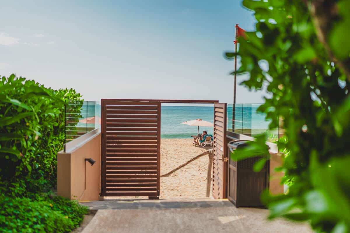 A wooden gate opens directly onto a sandy beach, offering a clear view of the turquoise ocean and a person relaxing under a beach umbrella in the distance. Lush green foliage frames the pathway to the gate.