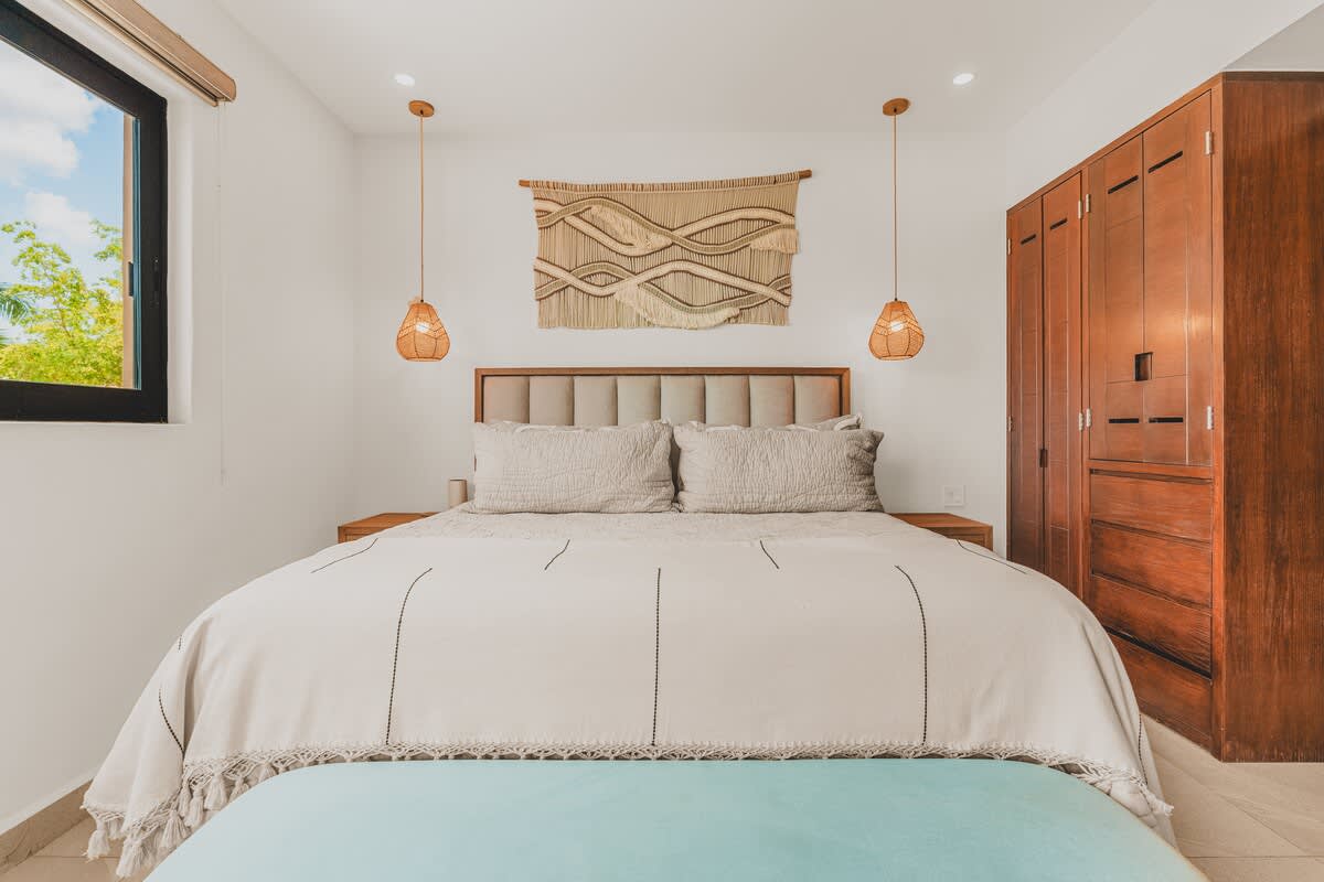 A bright bedroom showcases a large bed with a light duvet and pillows, a padded headboard, a light blue bench, and two woven pendant lights flanking a textured wall hanging. A dark wooden wardrobe is visible on the right.