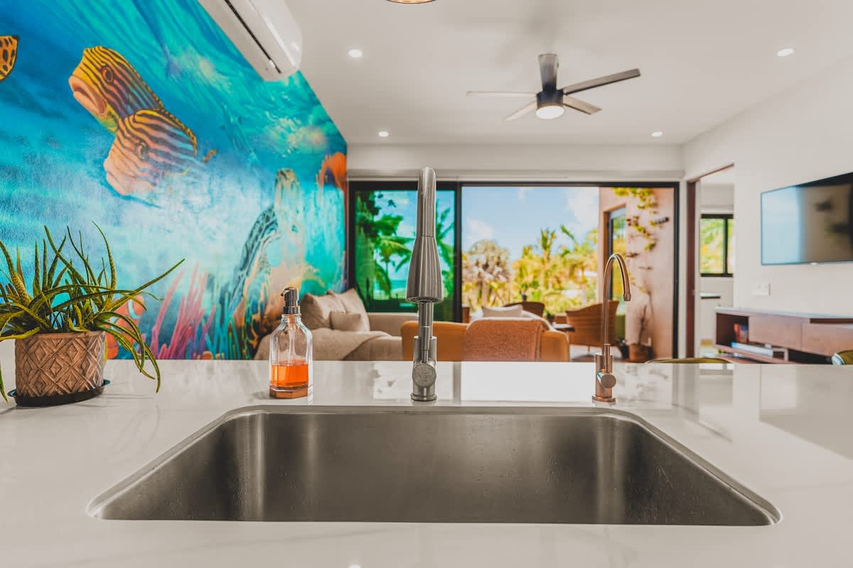 Close-up of the kitchen sink area on the island, showing a stainless steel basin and a modern pull-down faucet, with a plant and soap dispenser on the white countertop.