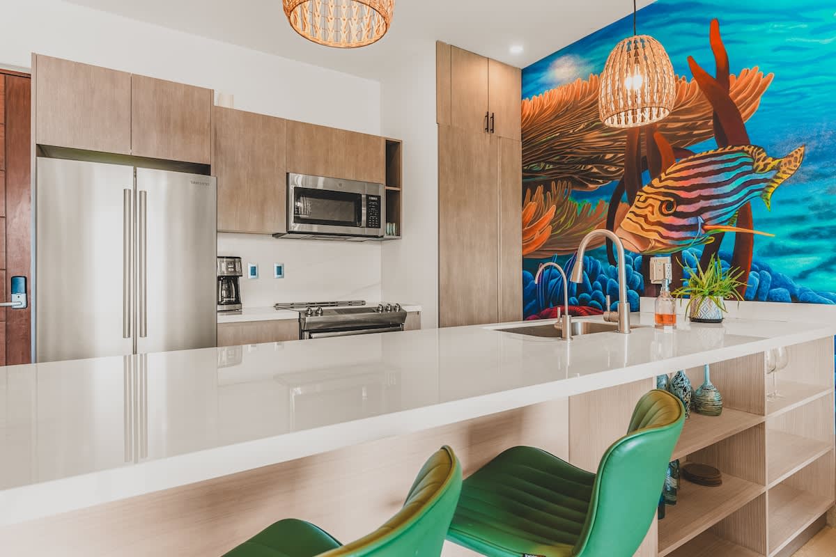 Modern kitchen with light wood cabinetry, white quartz countertops, and stainless steel appliances, featuring a vibrant underwater mural on the wall and two green bar stools at the island.