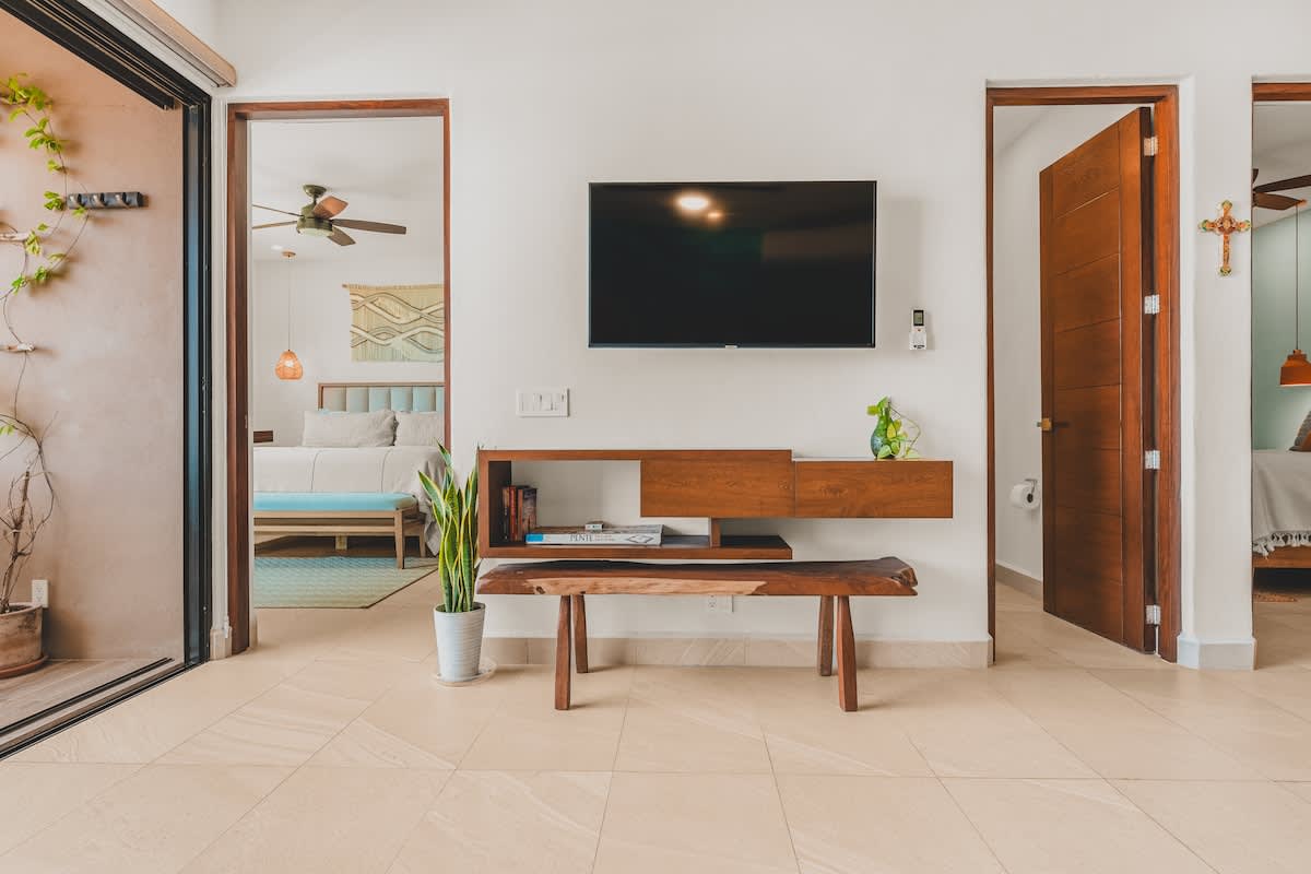 View of the living room area with a wall-mounted TV above a modern wooden console and bench, with open doorways leading to other rooms and a sliding glass door to the outside.