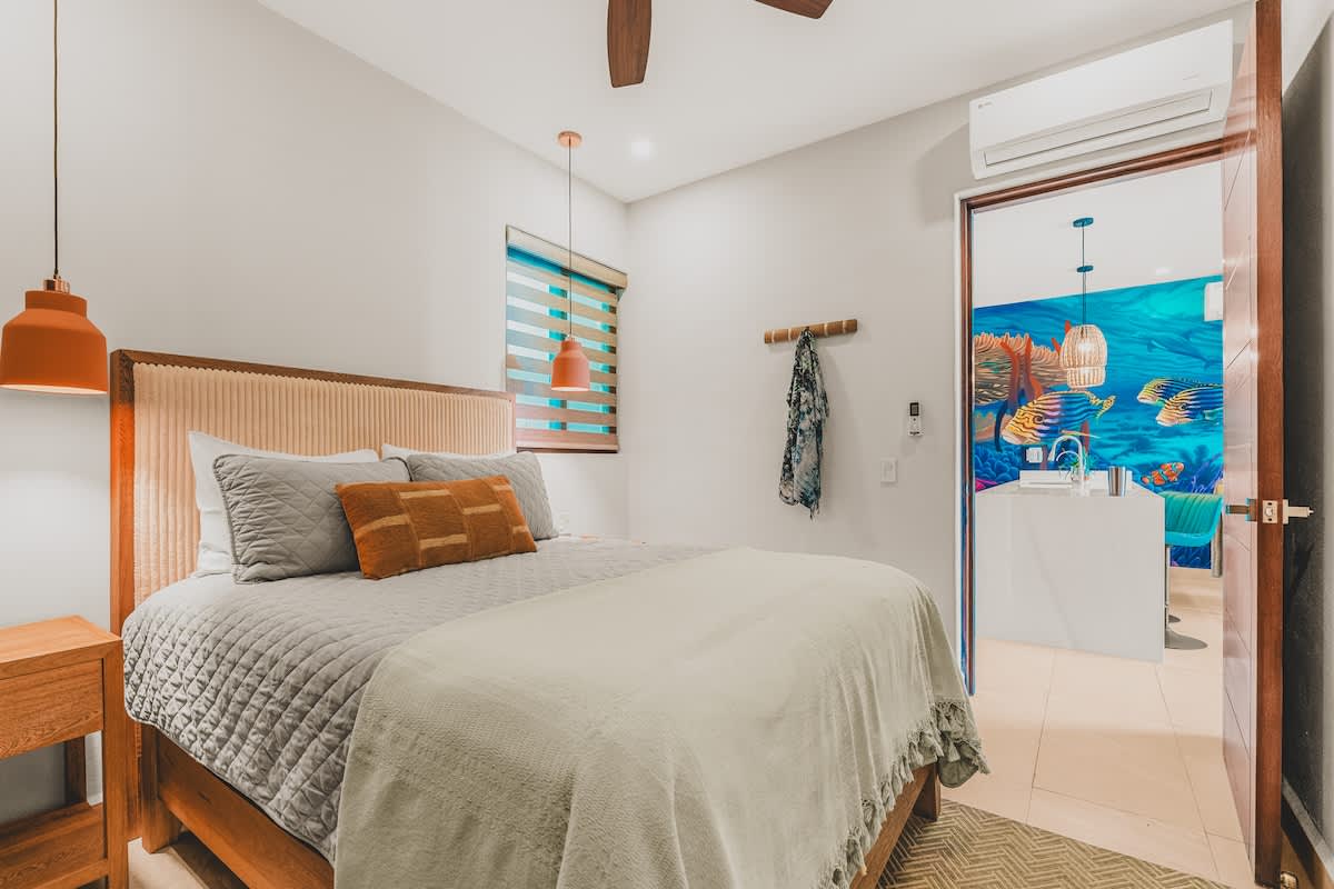 A bright bedroom features a queen-sized bed with a light beige upholstered headboard, wooden nightstands, and modern orange pendant lights. An open doorway reveals an ensuite bathroom with a vibrant underwater-themed mural, adding a playful touch.