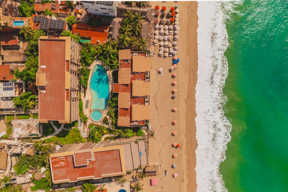 An aerial view showcases the beachfront property, featuring a large swimming pool, multiple buildings with terracotta roofs, and a wide sandy beach lined with colorful umbrellas next to the turquoise ocean.