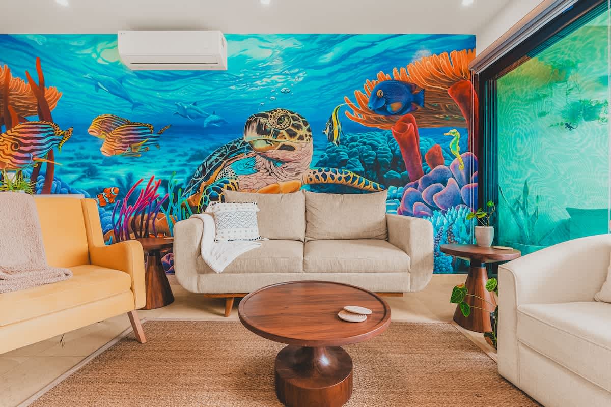 Living room with a beige sofa and a yellow armchair, centered around a round wooden coffee table, set against a colorful underwater mural and a natural fiber rug.