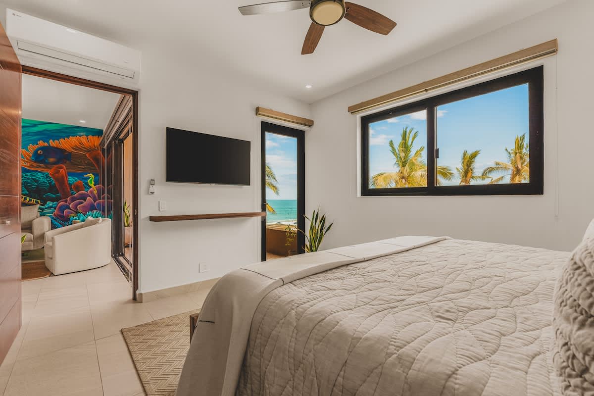 A bright bedroom with a large bed, a wall-mounted TV, and a window offering ocean views and palm trees. A doorway leads to an adjacent room with a vibrant underwater-themed mural.