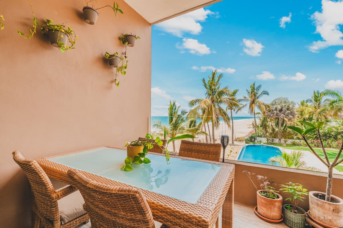 A spacious balcony features a glass-top rattan dining table with four chairs, overlooking a vibrant blue ocean, a swimming pool, and lush palm trees under a bright sky. Potted plants adorn the balcony wall and floor, adding a touch of greenery.