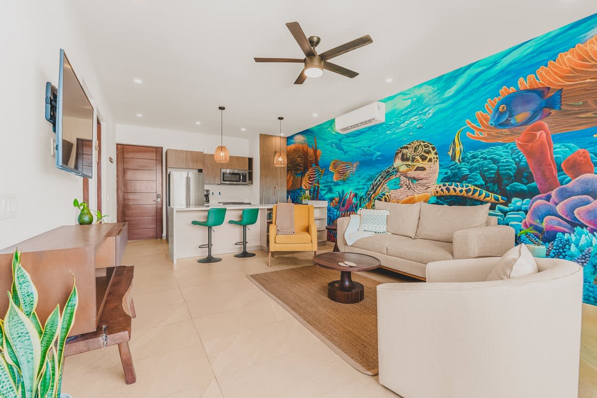 A modern open-concept living space features a vibrant underwater mural, a beige sectional sofa, a yellow accent chair, and a kitchen island with green bar stools. The room has light tile flooring and a ceiling fan.