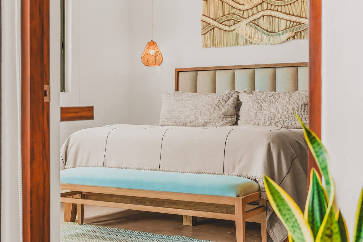 A cozy bedroom features a large bed with a light-colored duvet, a padded headboard, a light blue bench at the foot, and a woven wall hanging above the bed.