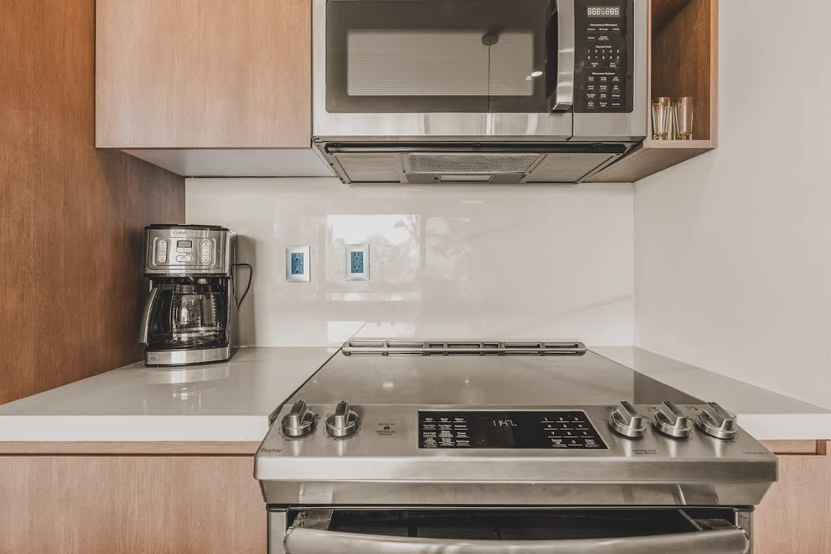 Close-up of the kitchen counter featuring a stainless steel range with a flat cooktop, a coffee maker, and a microwave oven mounted above, set against a white backsplash and light wood cabinets.