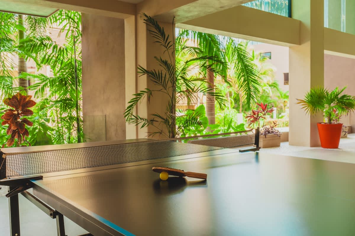 An indoor game area features a black ping pong table with a paddle and ball, set against a backdrop of lush tropical greenery visible through large open windows. The space has light-colored flooring.