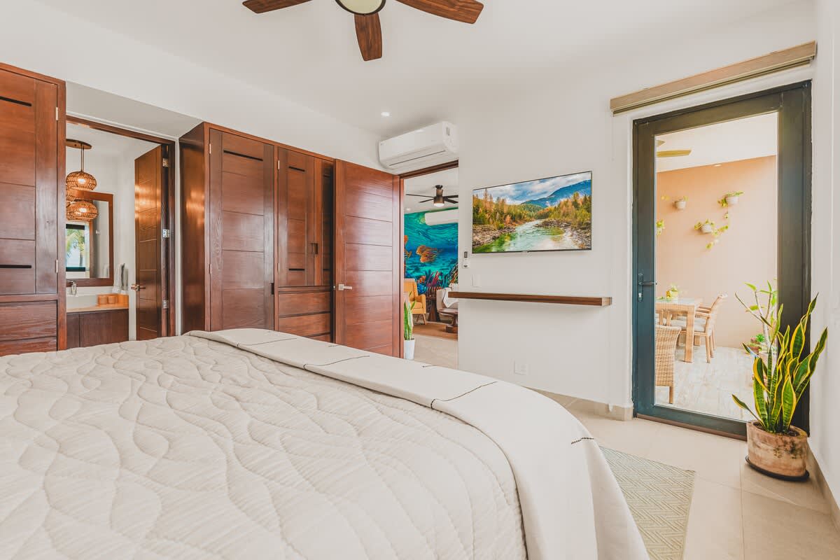 The bedroom features a large bed, a wall-mounted TV with a floating shelf, and extensive dark wooden built-in wardrobes. A doorway reveals a vibrant, colorful mural in an adjacent room.