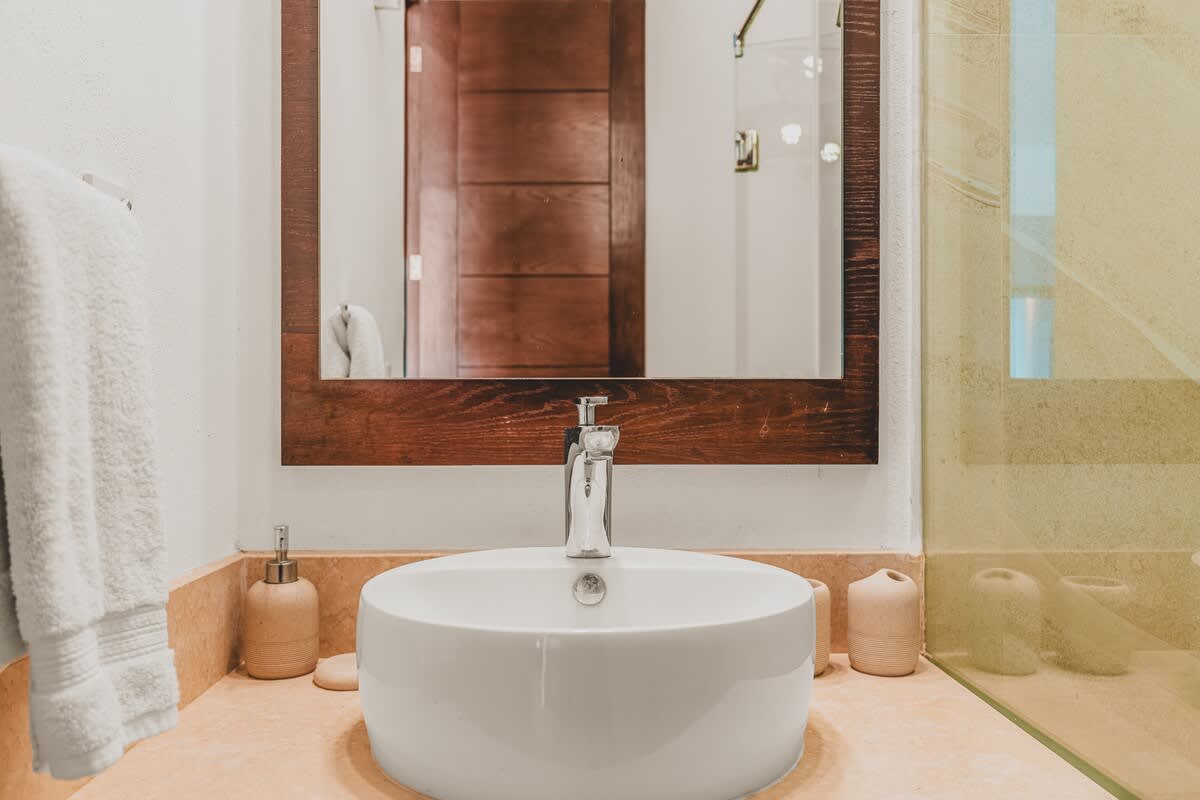 A modern bathroom features a white vessel sink on a light-colored countertop, a wooden-framed mirror, and a glass-enclosed shower with light-toned tiles.