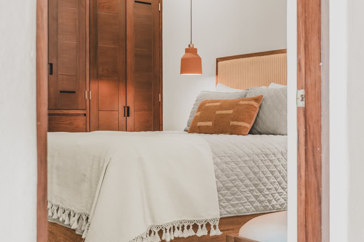 A view into bedroom 2 from the doorway highlights the queen-sized bed with its light beige upholstered headboard, flanked by wooden nightstands and modern orange pendant lights. Dark wooden built-in wardrobes are visible on the left, offering ample storage.