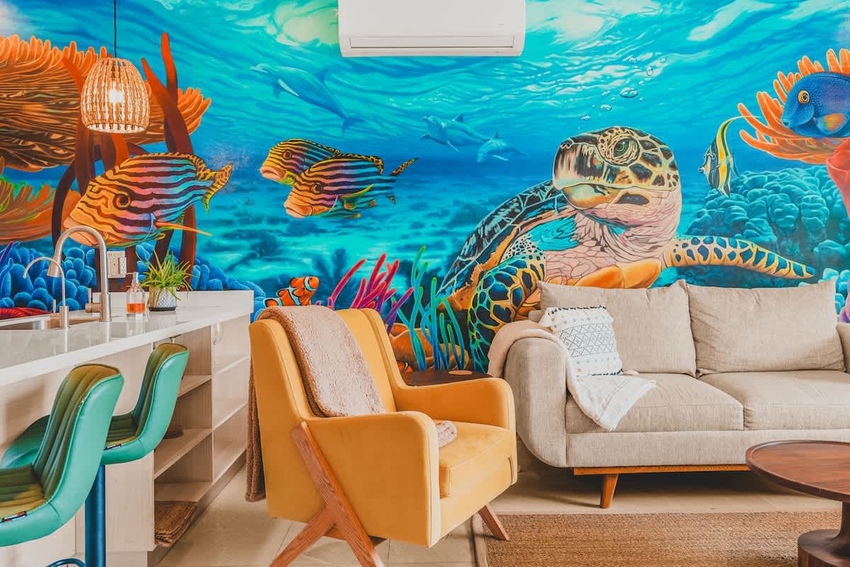 A vibrant living room features a large, colorful mural depicting an underwater scene with a sea turtle, fish, and coral, dominating the wall behind a light-colored sofa and a yellow armchair. The space is illuminated by a modern pendant light and natural light.
