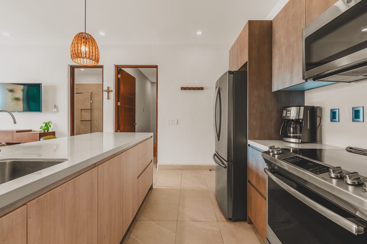 View of the kitchen showing light wood cabinets, white countertops, a stainless steel refrigerator, and a range with an overhead microwave, with two wooden doors visible in the background.