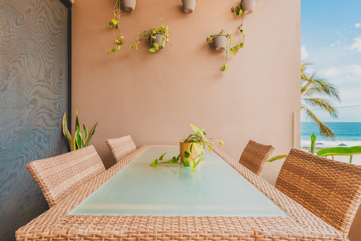 A well-appointed balcony dining area features a large glass-top rattan table with six chairs, set against a warm-toned wall adorned with hanging potted plants. The ocean and beach are visible in the distance, framed by lush palm trees.