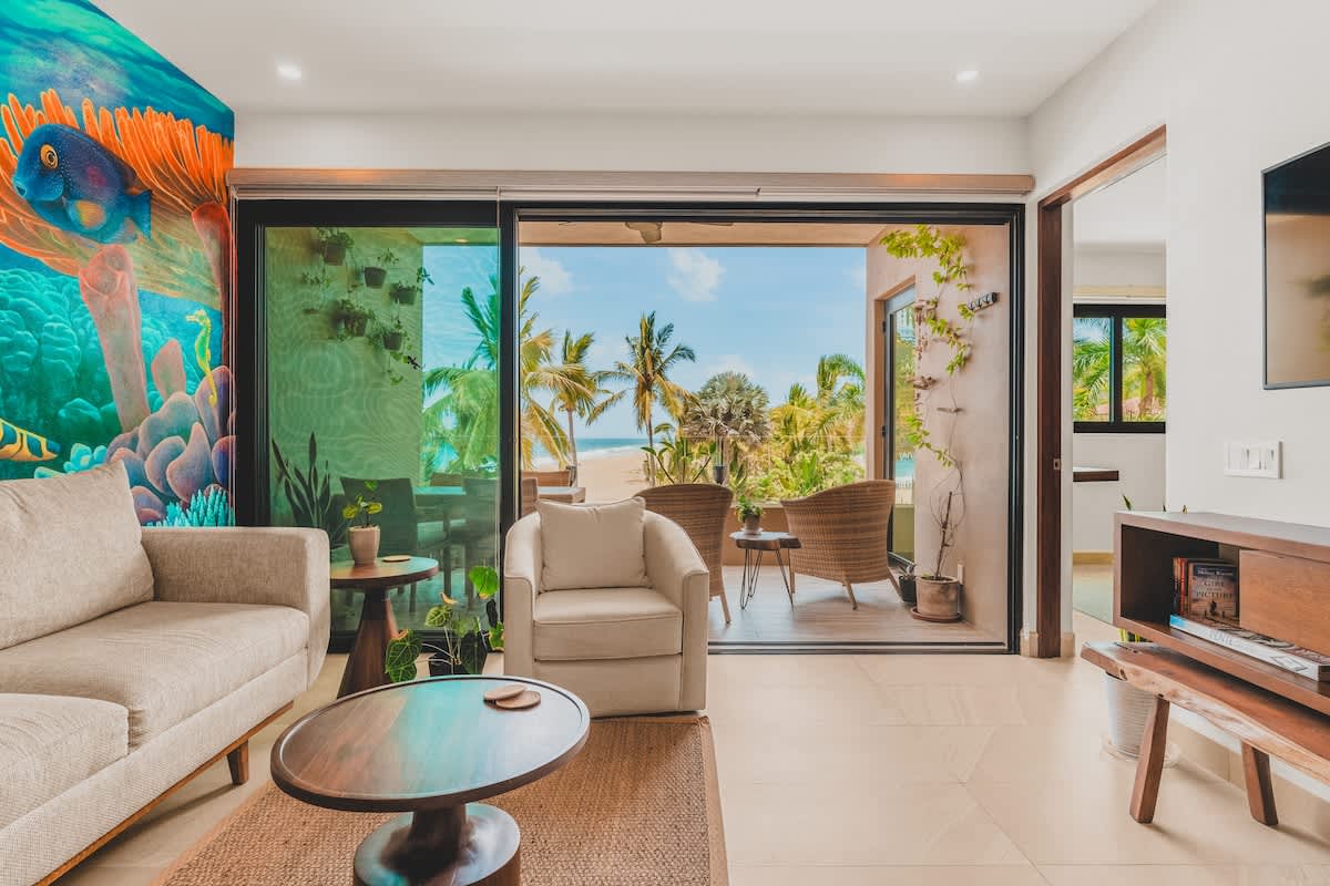 A bright living room with light tile floors and a large, colorful underwater-themed mural on the left wall, opens to a balcony with ocean and palm tree views through sliding glass doors. The room is furnished with a light-colored sofa, an armchair, and a round wooden coffee table.