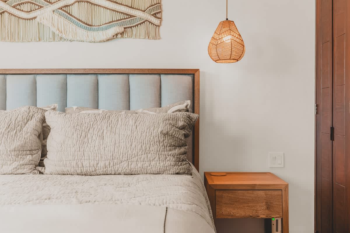 A close-up of the bedroom shows a padded headboard, light-colored pillows and duvet, a wooden nightstand with a light switch, and a woven pendant light.