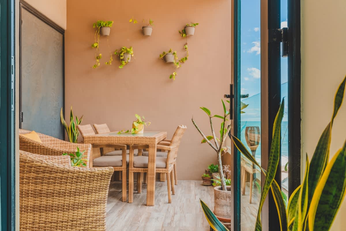 A close-up view of a balcony's outdoor dining area, featuring a glass-top rattan table and chairs, with a glimpse of the ocean and palm trees in the background. Various potted plants, including a tall snake plant, add natural elements to the space.