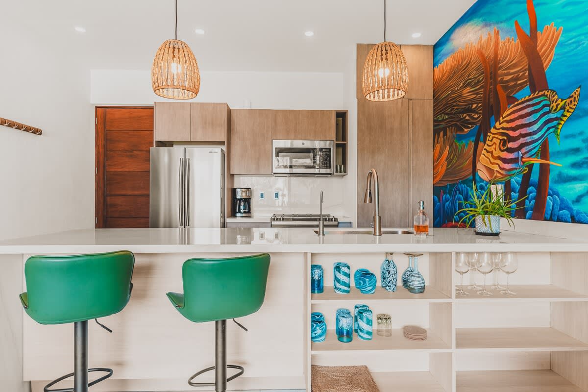 A modern kitchen features light wood cabinetry, stainless steel appliances, and a large island with a white countertop and two green bar stools. A vibrant underwater mural with fish and coral covers the wall, adding a unique artistic touch.