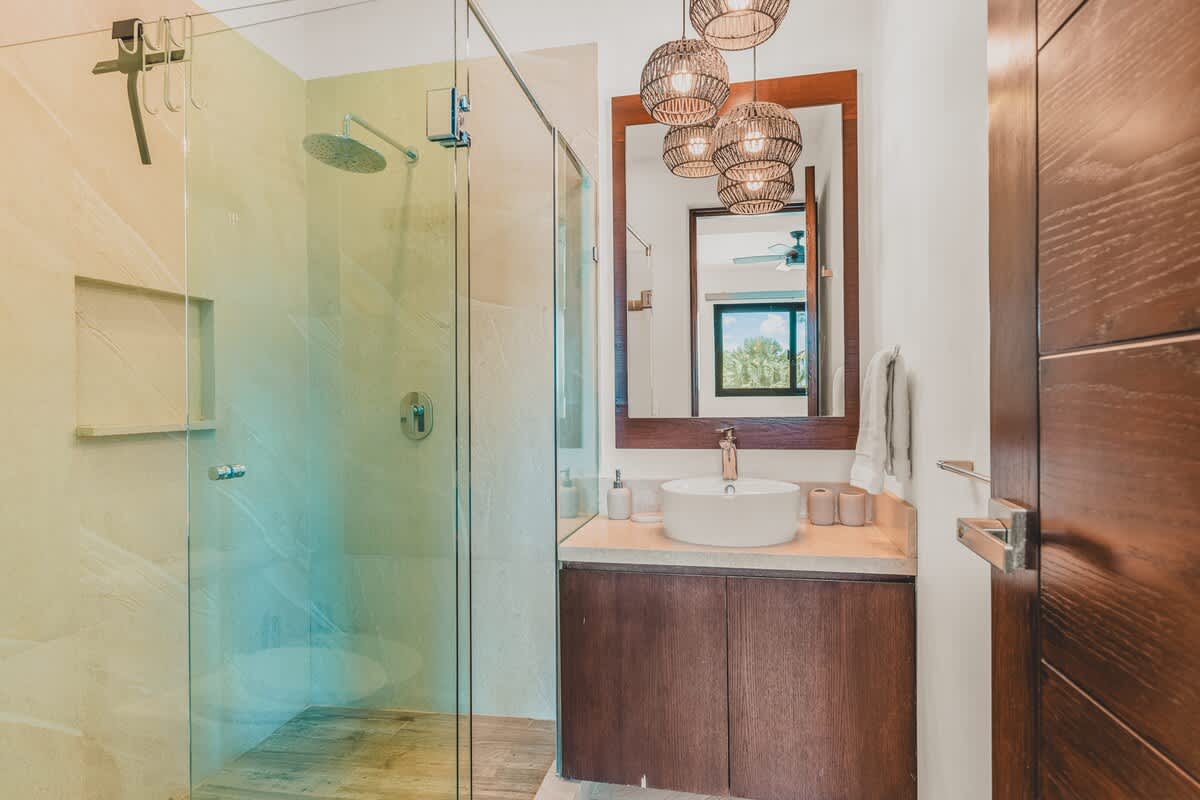 A contemporary bathroom features a glass-enclosed shower with a rain showerhead, a vanity with a vessel sink, a wooden-framed mirror, and three woven pendant lights.