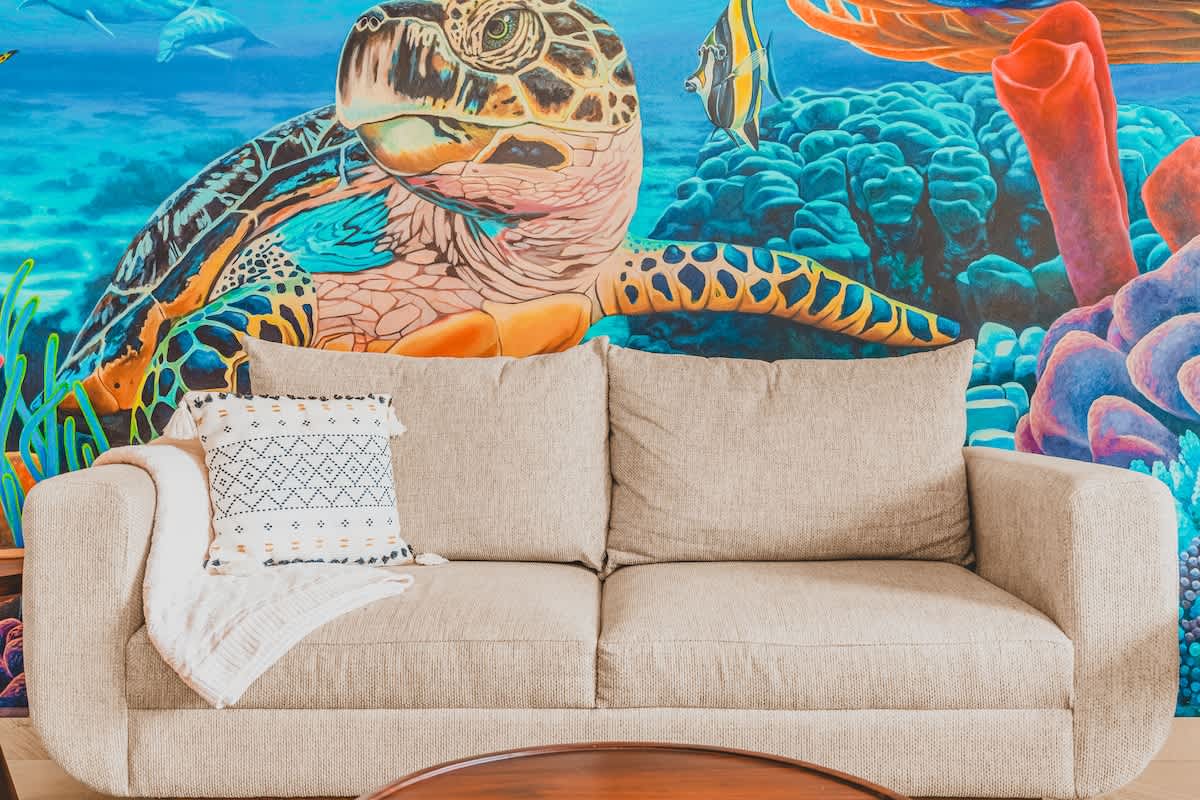Cozy living room featuring a light beige sofa with a patterned throw pillow against a vibrant underwater mural, with a round wooden coffee table in the foreground.