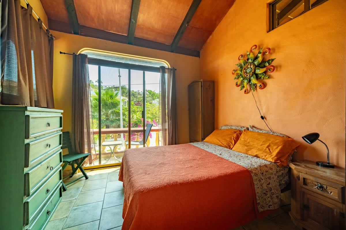 A cheerful orange bedroom features a queen bed with matching bedding, a green dresser, and a large window offering balcony access and views of lush greenery. The room boasts a vaulted wooden ceiling and tiled floors.