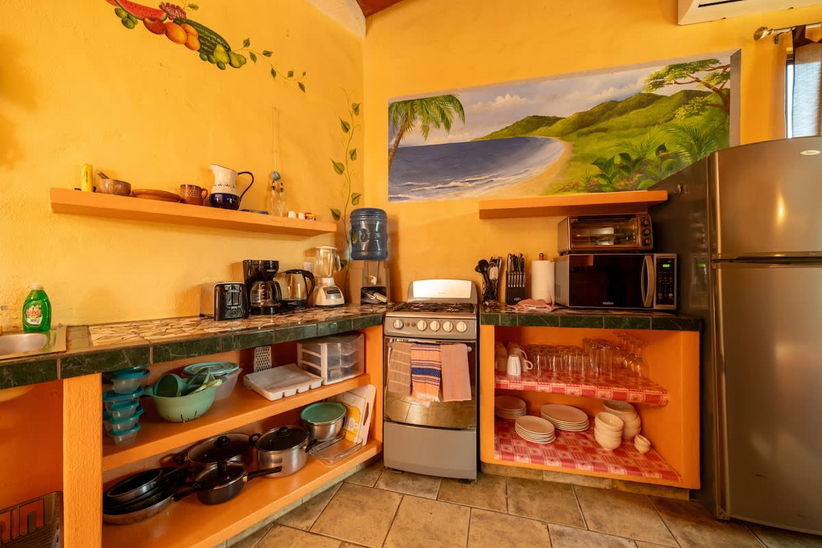A well-equipped kitchen with yellow walls and tiled countertops, featuring a refrigerator, gas stove, microwave, and various small appliances. Open shelves hold dishes and cooking essentials, with a beach mural above.