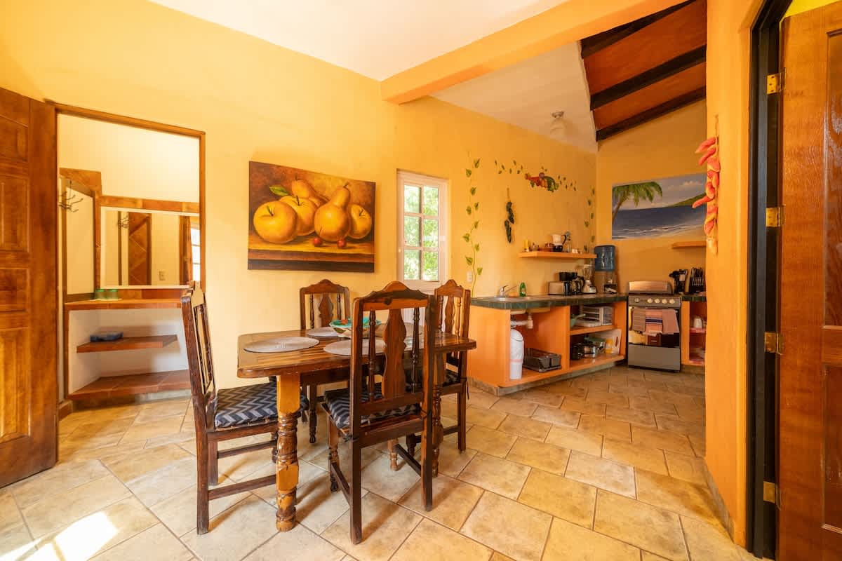 An open-concept dining and kitchen area with warm yellow walls and tiled floors. A wooden dining table is central, with the kitchen visible in the background featuring a stove, sink, and open shelving.