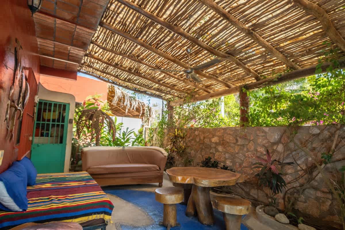 A spacious and shaded outdoor patio area with a natural wood slat roof, offering various seating options including a daybed with colorful blankets and a brown sofa, alongside a large rustic wooden table and stools. The area is surrounded by lush tropical foliage and a stone wall.