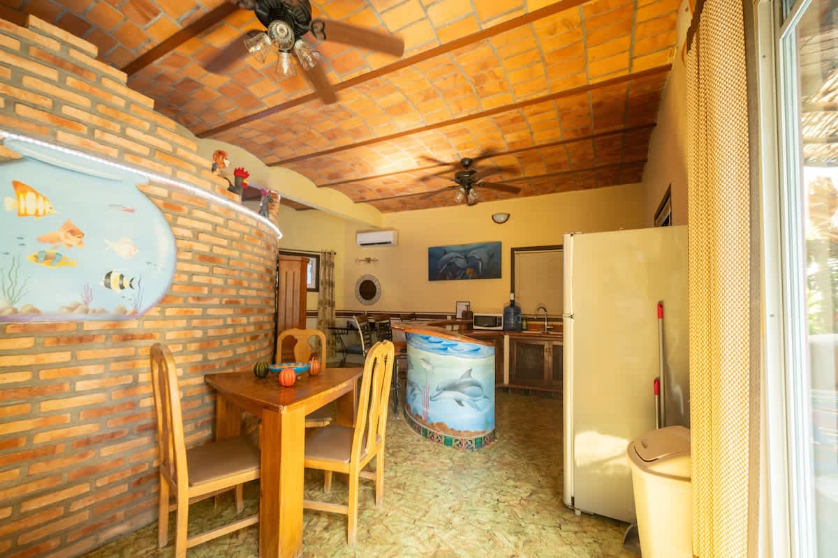 A bright kitchen and dining space with a small wooden dining table and chairs, adjacent to a brick wall with a fishbowl mural. The kitchen area features a refrigerator, a counter with a dolphin mural, and a vaulted terracotta tile ceiling, with natural light from a large sliding door.
