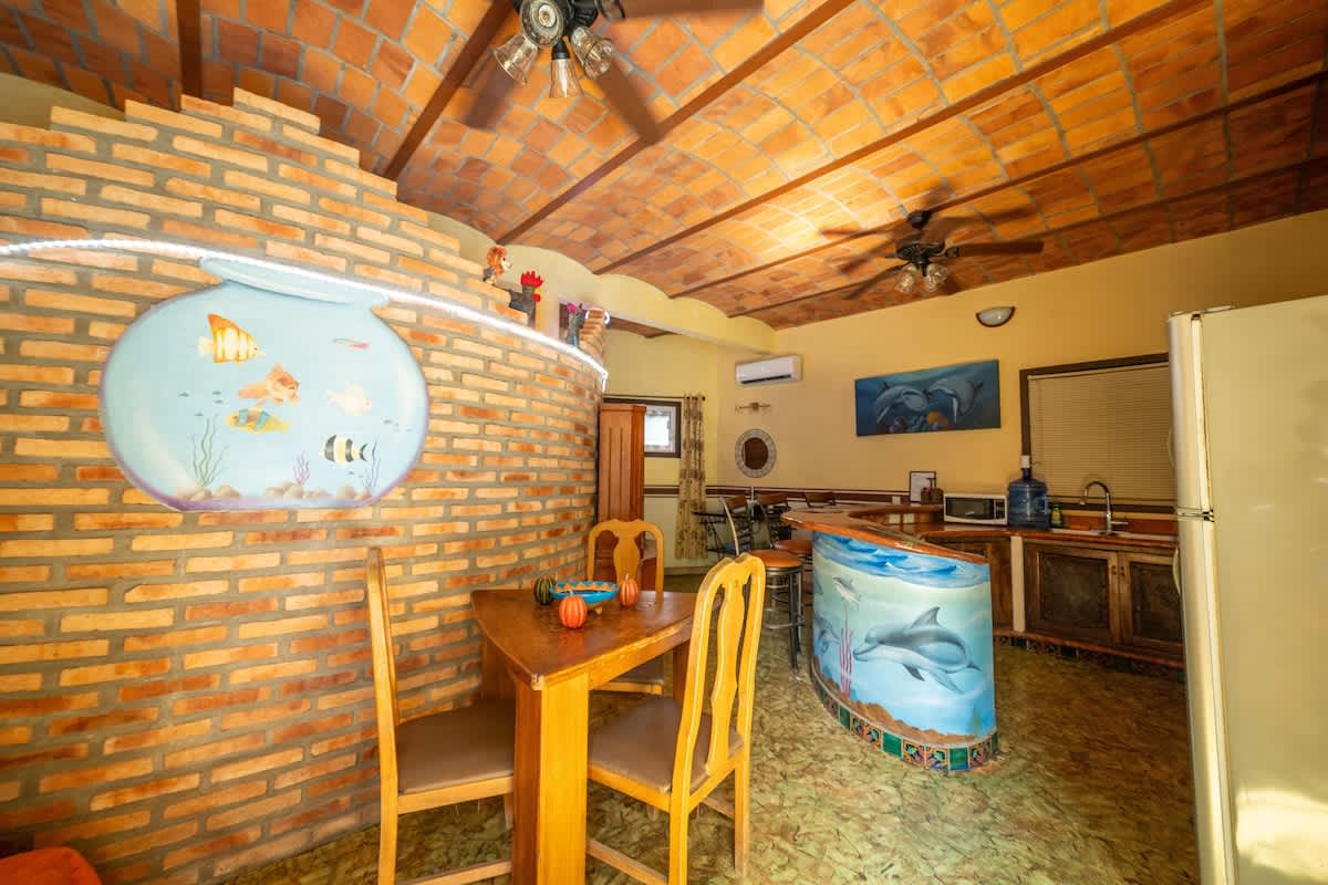 An open-concept kitchen and dining area features a small wooden dining table with two chairs next to a brick accent wall with a fishbowl mural. The space includes a kitchen counter with a dolphin mural, a refrigerator, and a vaulted terracotta tile ceiling with ceiling fans.