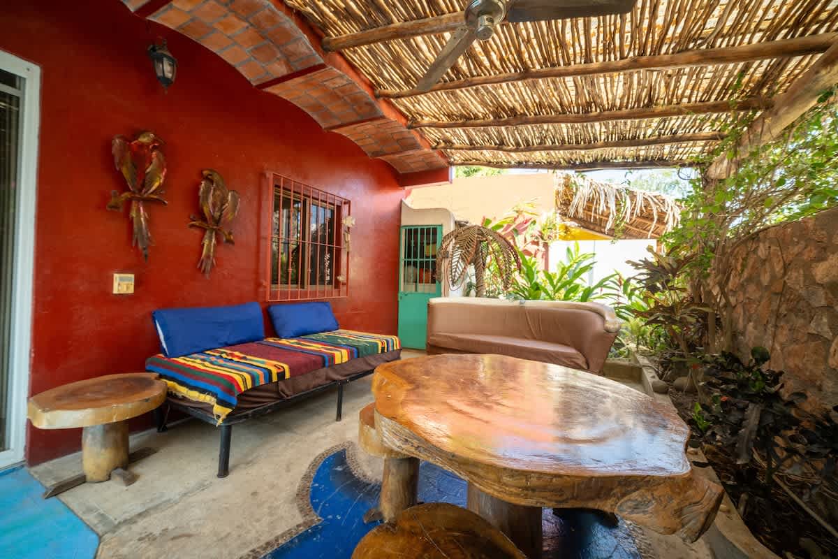 An inviting outdoor patio with a natural wood slat roof, featuring a daybed with colorful textiles and blue pillows, and a large rustic wooden table with stools. The red-painted wall is adorned with bird sculptures, and the area is enclosed by a stone wall and lush plants.