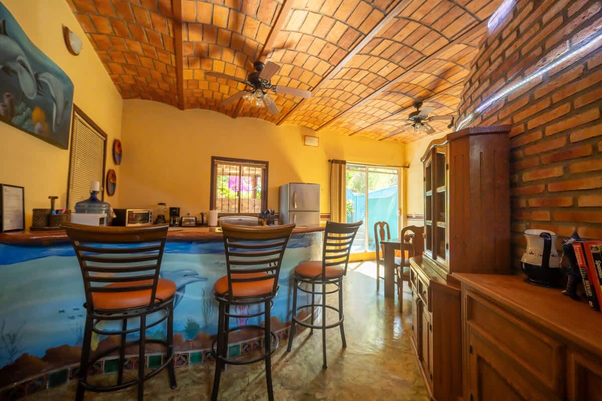 A spacious kitchen and bar area featuring a long counter with a dolphin mural and three bar stools, alongside a wooden hutch. The room has light yellow walls, a vaulted terracotta tile ceiling with ceiling fans, and a refrigerator in the background.