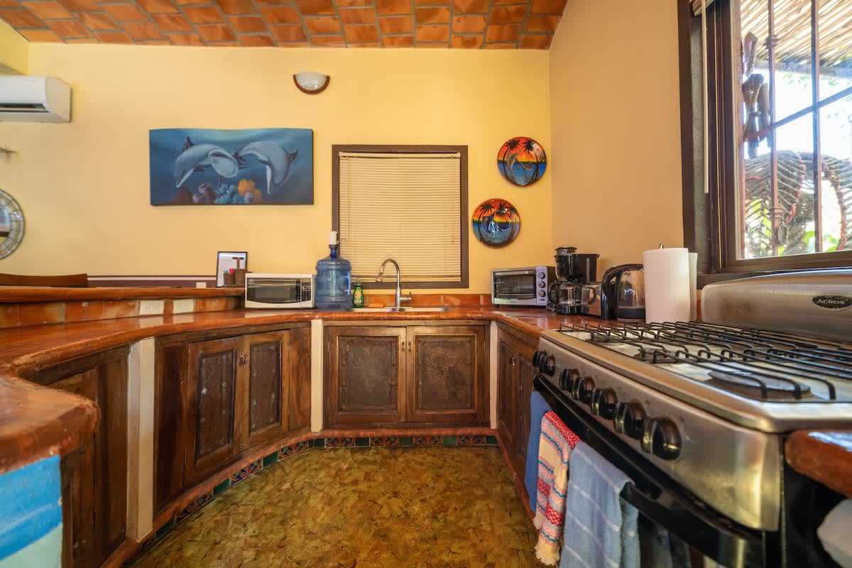 A well-equipped kitchen counter with rustic wooden cabinetry, a gas range, microwave, coffee maker, and toaster. The counter is topped with terracotta tiles, and a window with blinds provides natural light, complemented by decorative wall plates.