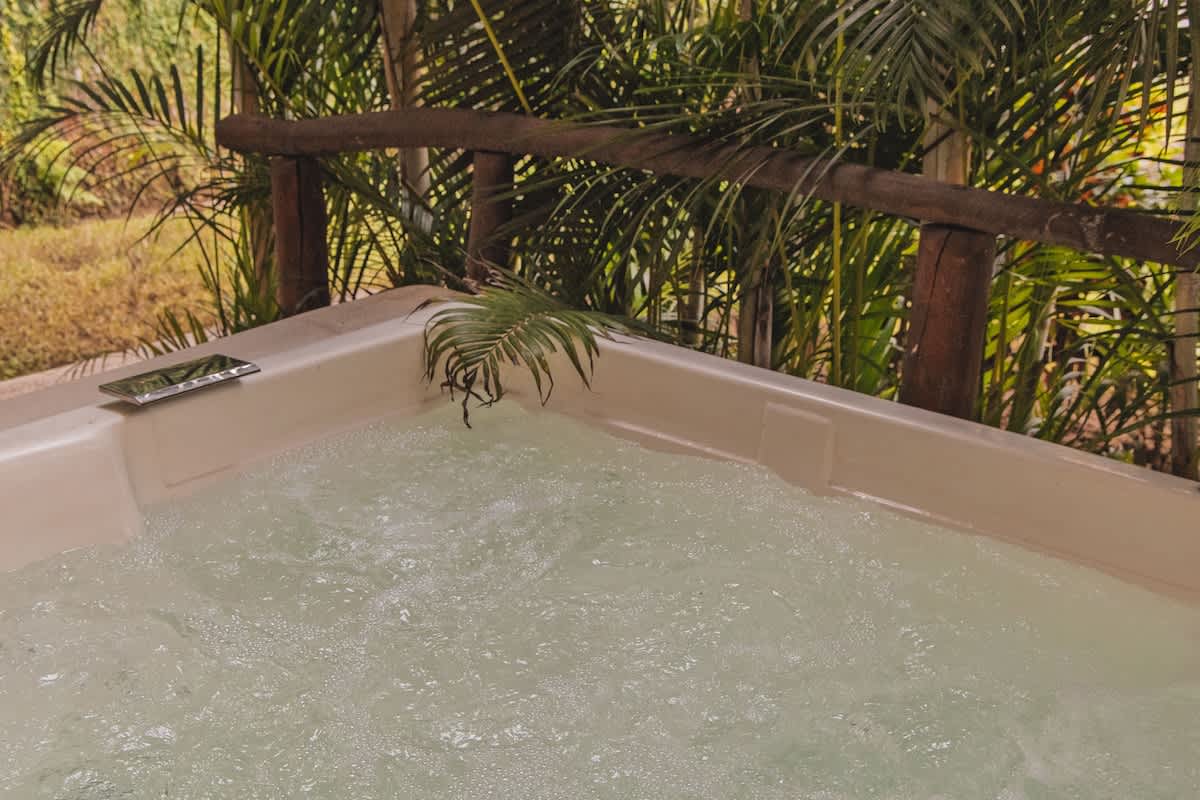 A close-up view of a bubbling hot tub with light-colored water, set against a backdrop of lush green palm fronds and a rustic wooden railing. The hot tub has a light beige or off-white finish.
