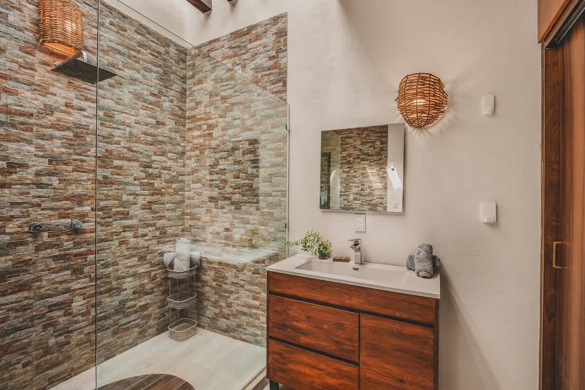 A close-up view of the bathroom vanity with a white countertop and sink, a mirror reflecting the shower, and a woven light fixture, adjacent to a glass-enclosed shower with a stacked stone wall.