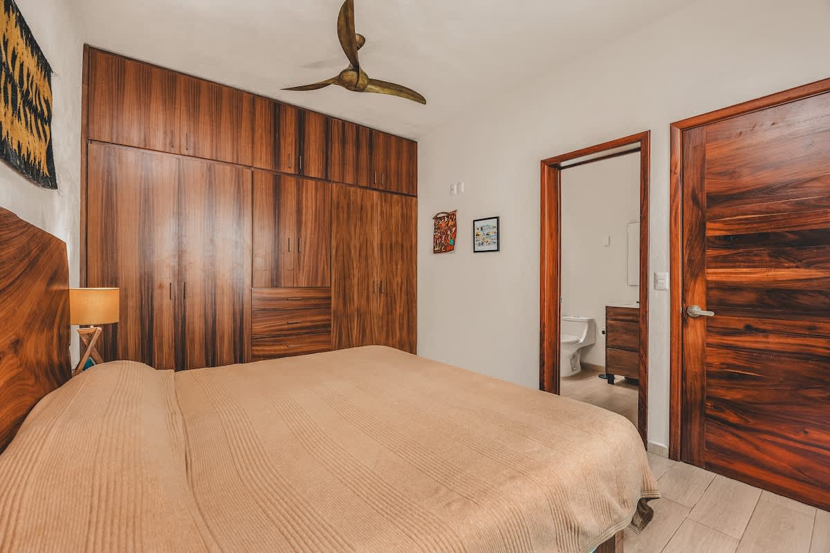 A warm bedroom features a large bed with a live-edge wooden headboard, a full wall of dark wood built-in wardrobes, and a blue rug, with a wooden door leading to an en-suite bathroom.