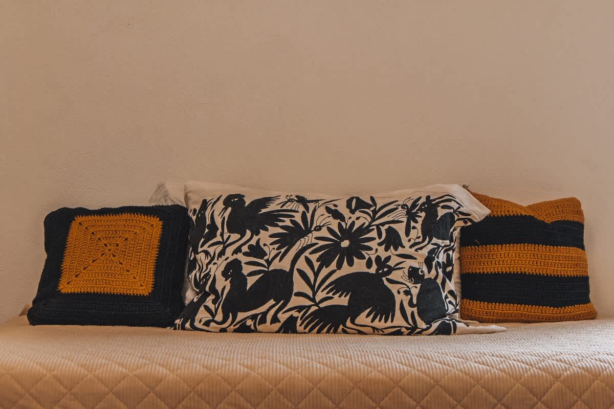 A bed is adorned with decorative pillows, including a central white pillow with black bird and floral patterns, flanked by two black and yellow crocheted square pillows, resting on a quilted bedspread.