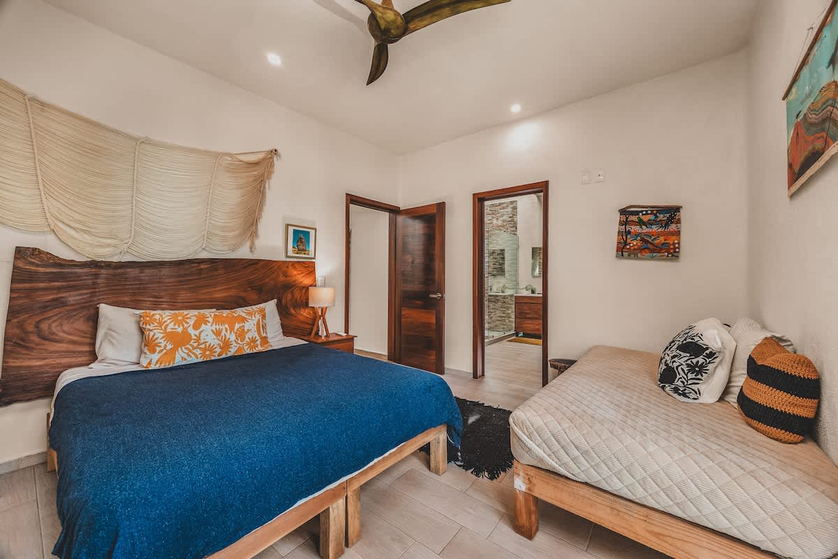 Spacious bedroom featuring a large bed with a unique live-edge wooden headboard, a daybed, and a doorway leading to an en-suite bathroom, complemented by a ceiling fan and bedside table with a lamp.