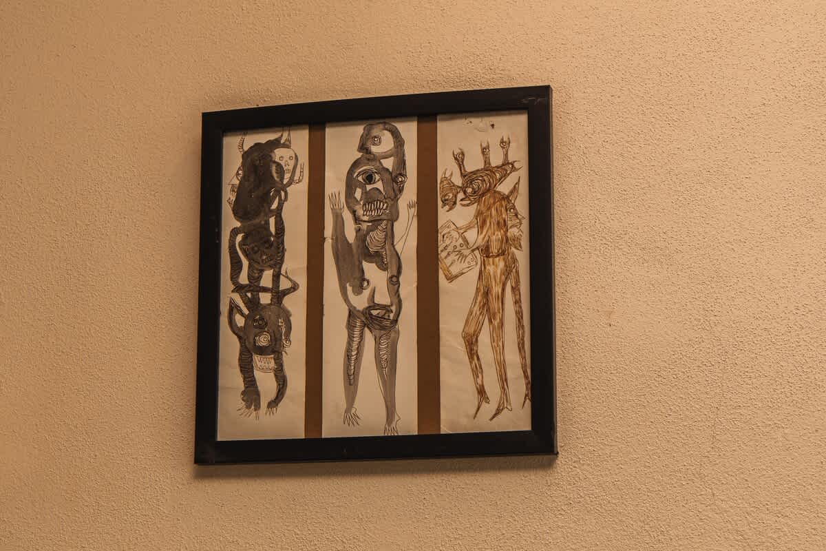 A framed piece of art featuring three abstract, dark-toned figures with intricate details hangs on a textured light brown wall, adding a unique artistic touch to the space.