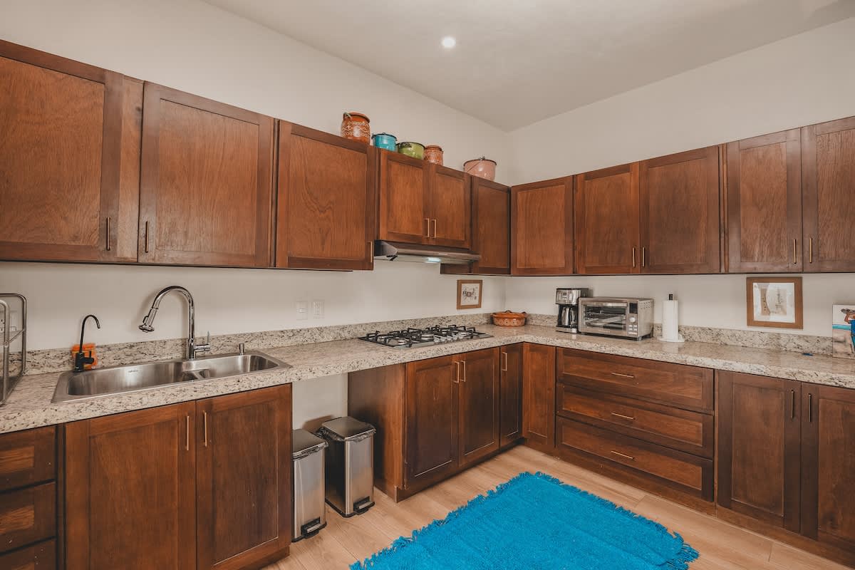 A well-equipped kitchen showcases dark wood cabinetry, speckled granite countertops, a gas stovetop, and a double sink. Stainless steel trash cans sit on a light wood floor, and a blue textured rug adds a vibrant accent.