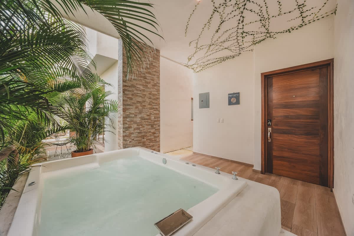 A wide view of the outdoor patio features a white built-in hot tub in the foreground, with a stone-tiled accent wall and a wooden entrance door visible in the background, all under a ceiling adorned with green vines.