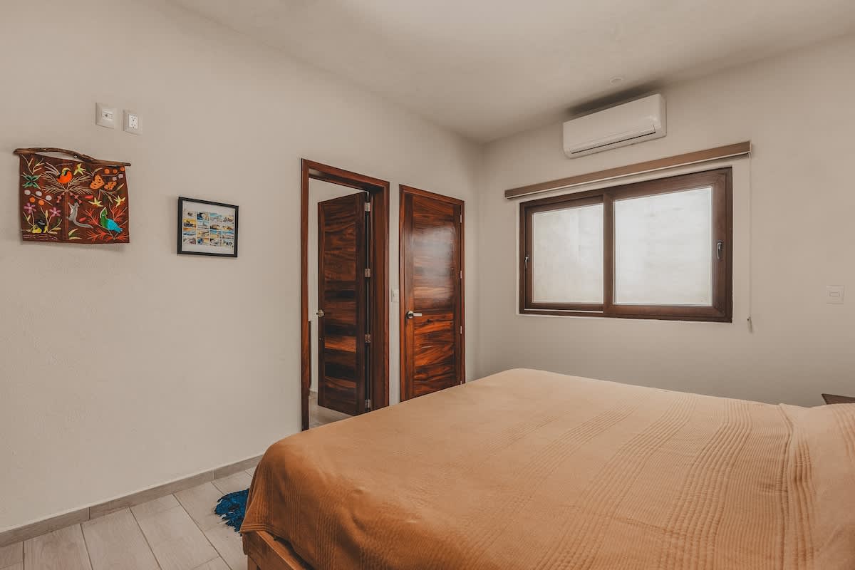 Bright bedroom with a large bed covered in a light brown blanket, featuring a wooden window with blinds, an air conditioning unit, and two dark wooden doors leading to other rooms. Artwork hangs on the wall.