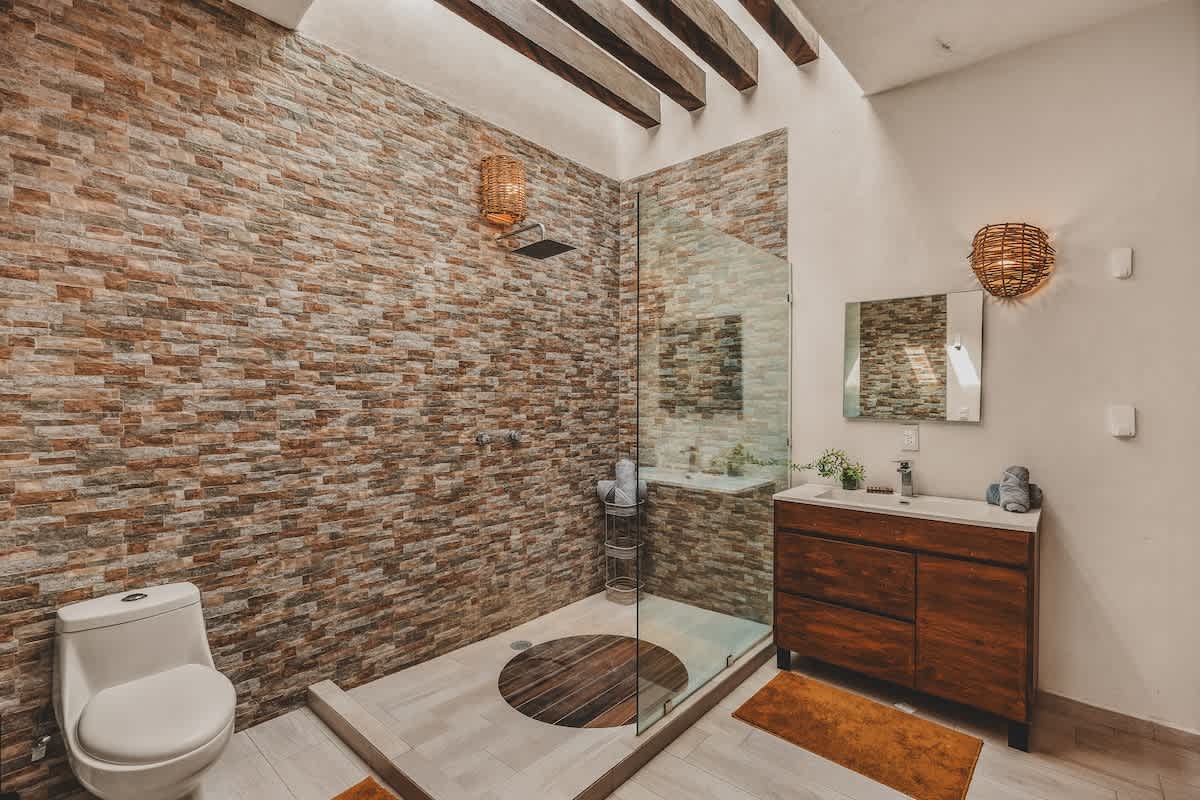 A modern bathroom with a spacious walk-in shower featuring a stacked stone accent wall and a glass enclosure, alongside a dark wood vanity with a white sink and mirror. Exposed wooden beams adorn the ceiling.